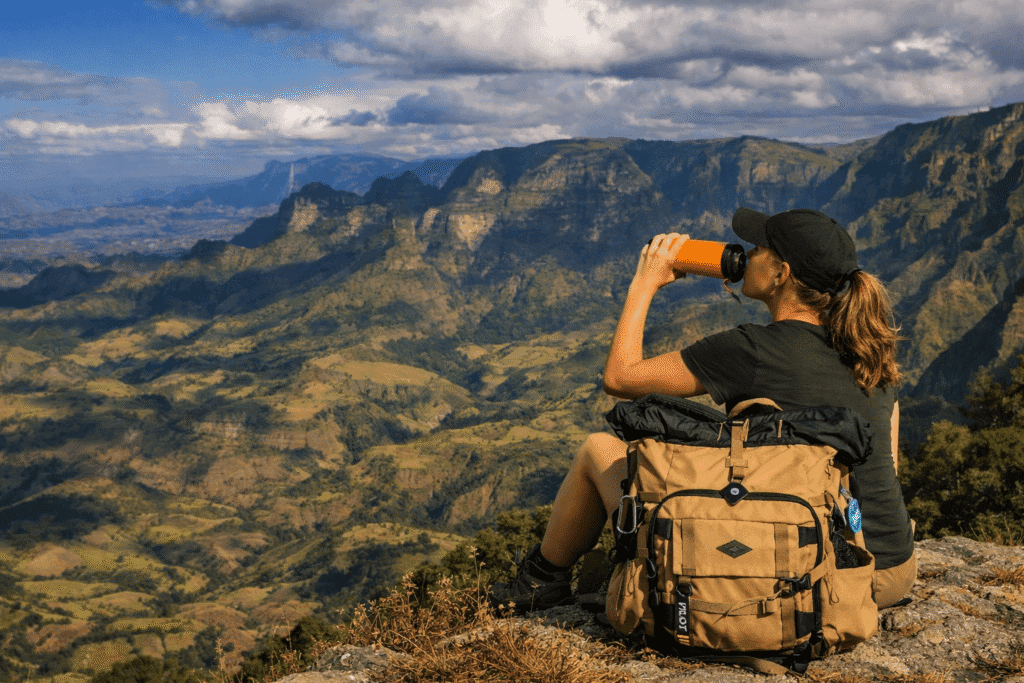 a female hiker sitting on a cliff edge, drinking from a water bottle and overlooking a dramatic Simien mountain landscape with deep valleys and rugged peaks.