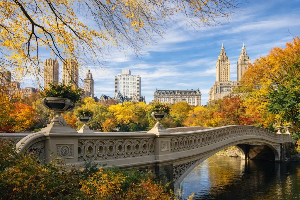 Bow Bridge arches over calm water in Central Park during peak fall foliage, with golden leaves framing the scene and the San Remo towers rising in the Manhattan skyline.